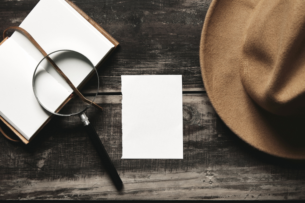 mysterious-detective-game-concept-opened-notebook-leather-cover-sheet-white-paper-felt-brown-hat-big-vintage-magnifier-steel-glasses-isolated-black-aged-wood-table-top-view _1_.jpg
