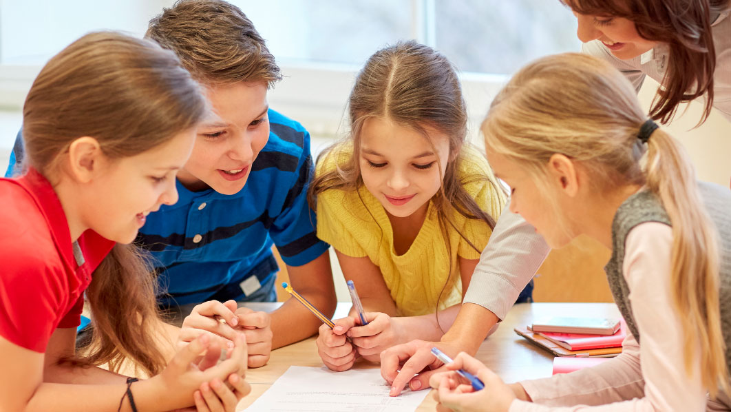 3 enfants avec une maitresse à l'école en train de lire un devoir 